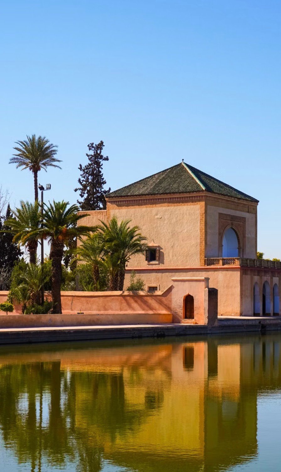 Pavillon historique des Jardins de la Ménara à Marrakech avec son bassin d’eau, ses palmiers et le reflet du bâtiment sous un ciel bleu.
